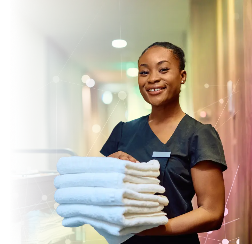 Smiling woman in black uniform holding a stack of folded white towels in a hallway.