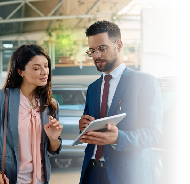 A man in a suit shows a tablet to a woman in a pink blouse and gray jacket in a car showroom.