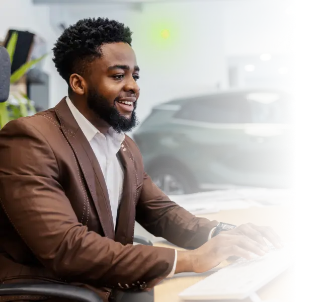 Smiling man in brown suit typing on a keyboard with a car blurred in the background.