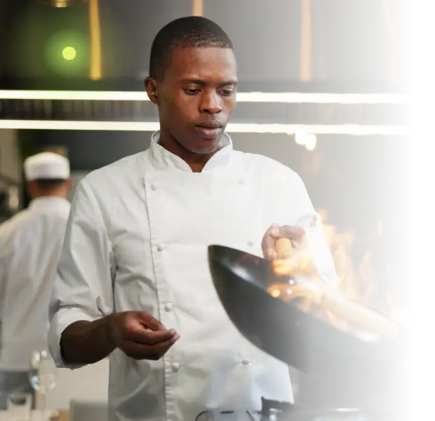 Chef in white coat cooking with flames in a wok in a professional kitchen.