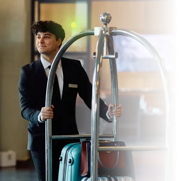 Hotel bellhop in suit pushing luggage cart with suitcases and bag in a lobby.