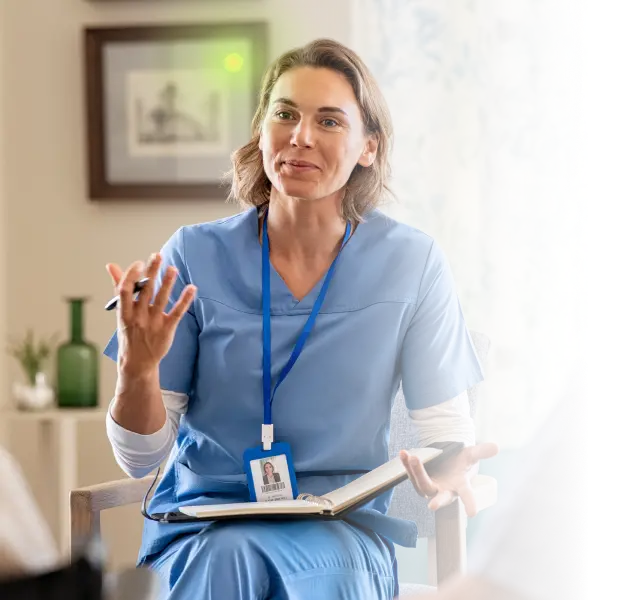 Female healthcare professional in blue scrubs talking and holding a notebook during a consultation.