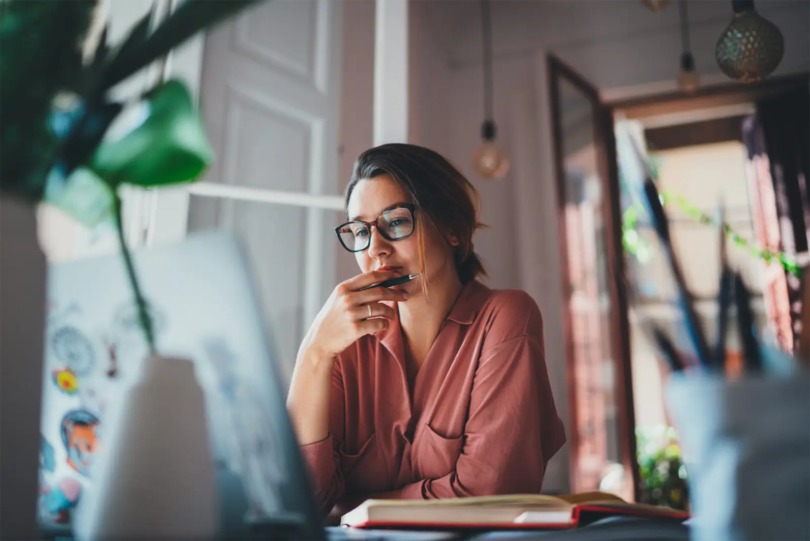 Woman with glasses, holding pen to mouth, focused on laptop in a cozy, well-lit room.