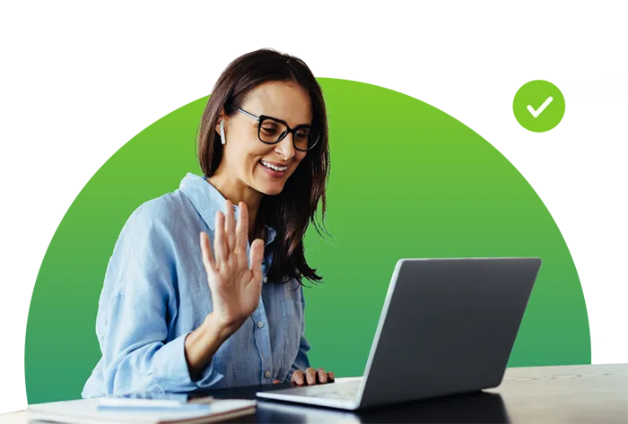 Smiling woman wearing glasses and earbuds waves while video calling on laptop against green background.