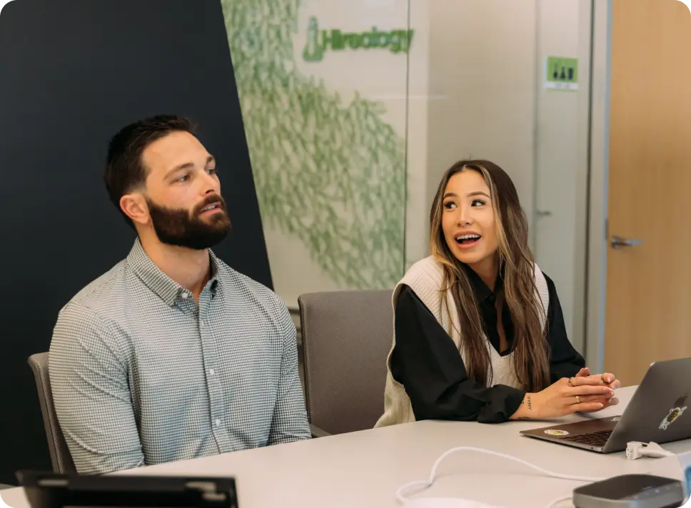 Man and woman seated at a table in an office during a meeting, woman speaking and smiling.