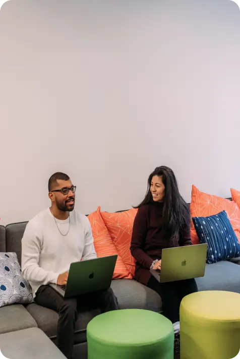 Man and woman sitting on couch with laptops, smiling and talking in a casual office space.