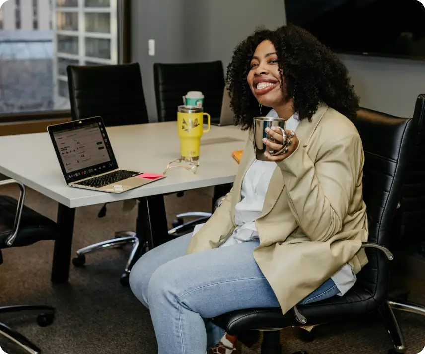 Smiling woman in blazer holding a silver mug while sitting in an office chair by a table with laptop.