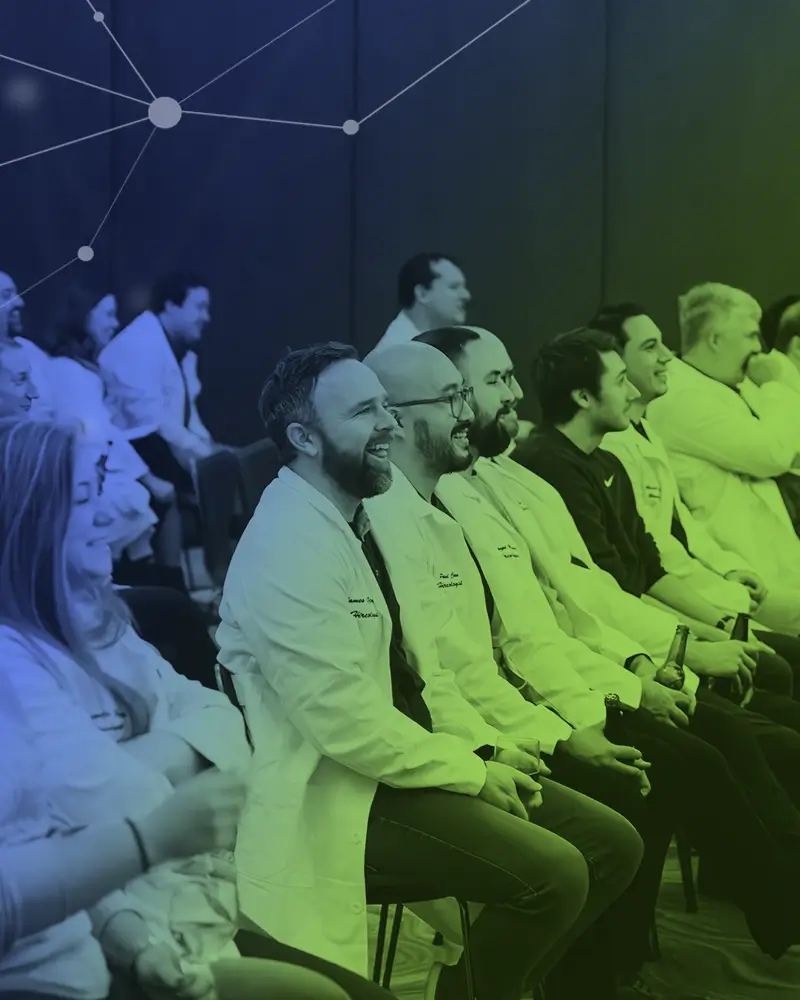 Group of people in white lab coats sitting in chairs, smiling and holding drinks, in a conference room.