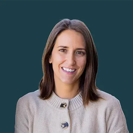 Smiling woman with straight brown hair wearing a beige buttoned sweater against teal background.
