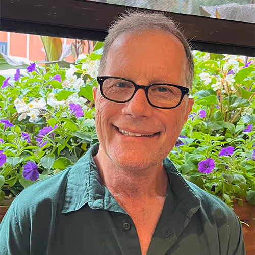 Smiling older man with glasses wearing a green shirt, standing in front of a window with flowers outside.