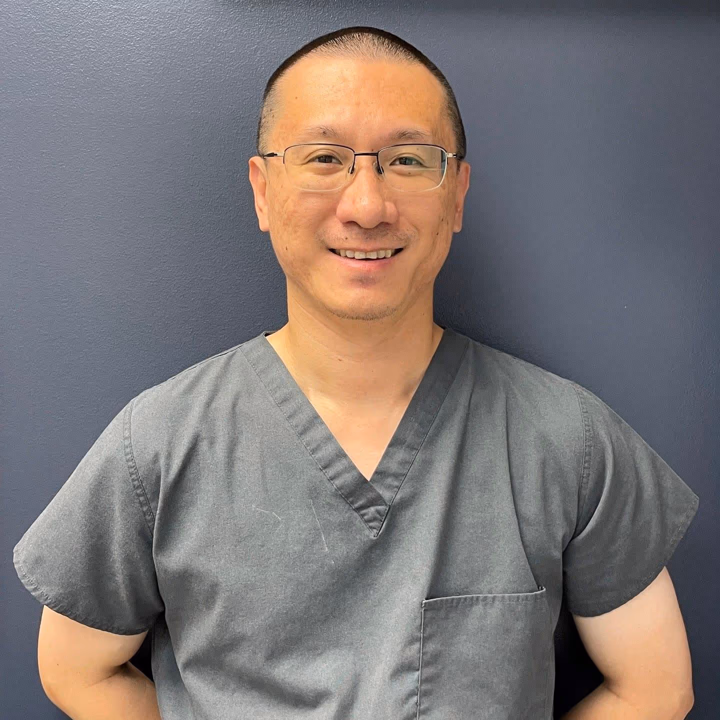 Smiling man wearing glasses and gray medical scrubs against a dark blue wall.