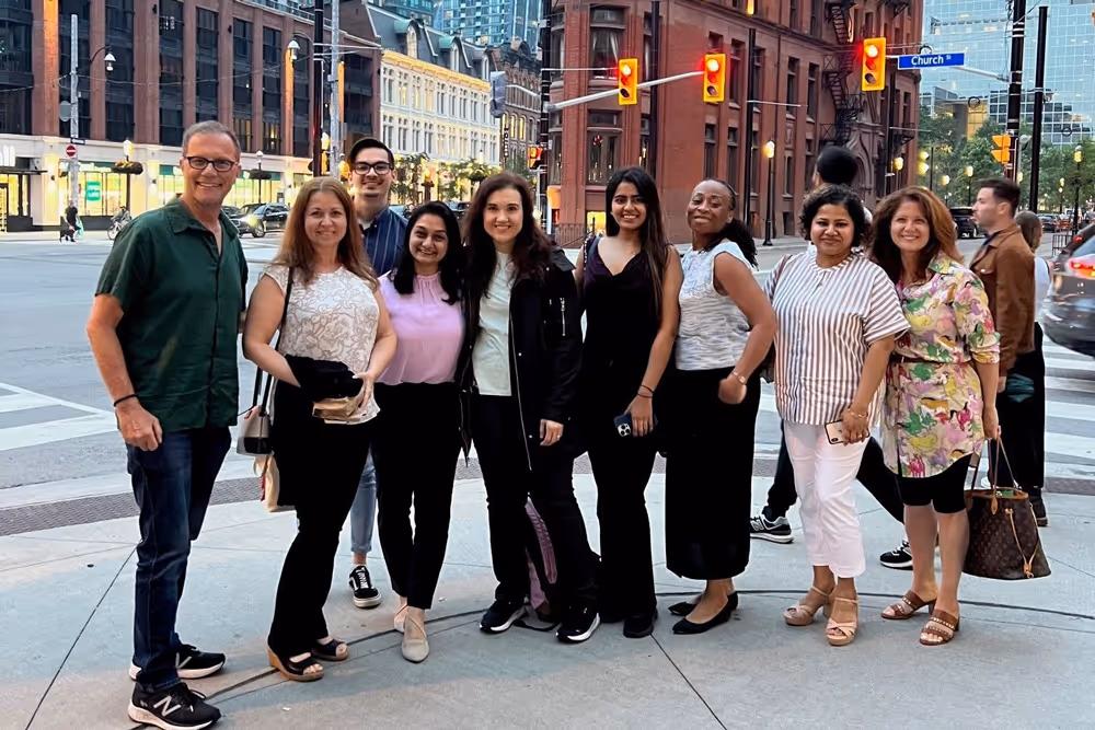 Group of nine smiling adults standing on a city street corner with buildings and traffic lights behind them.