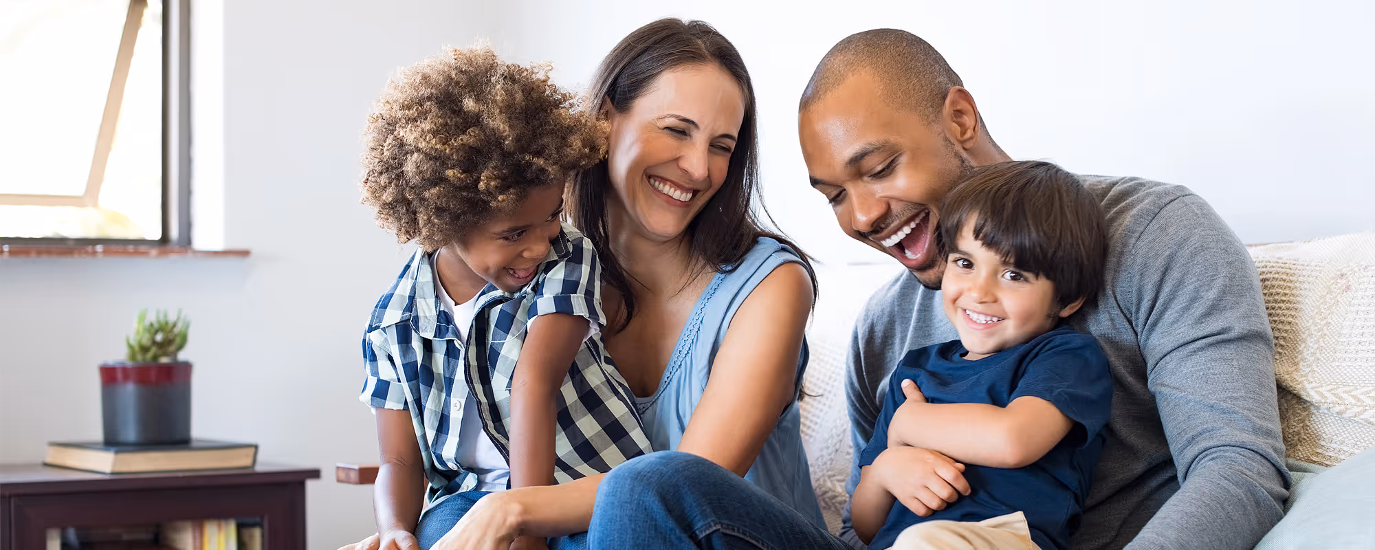 Smiling family of four sitting closely, with parents and two children sharing a happy moment on a couch.