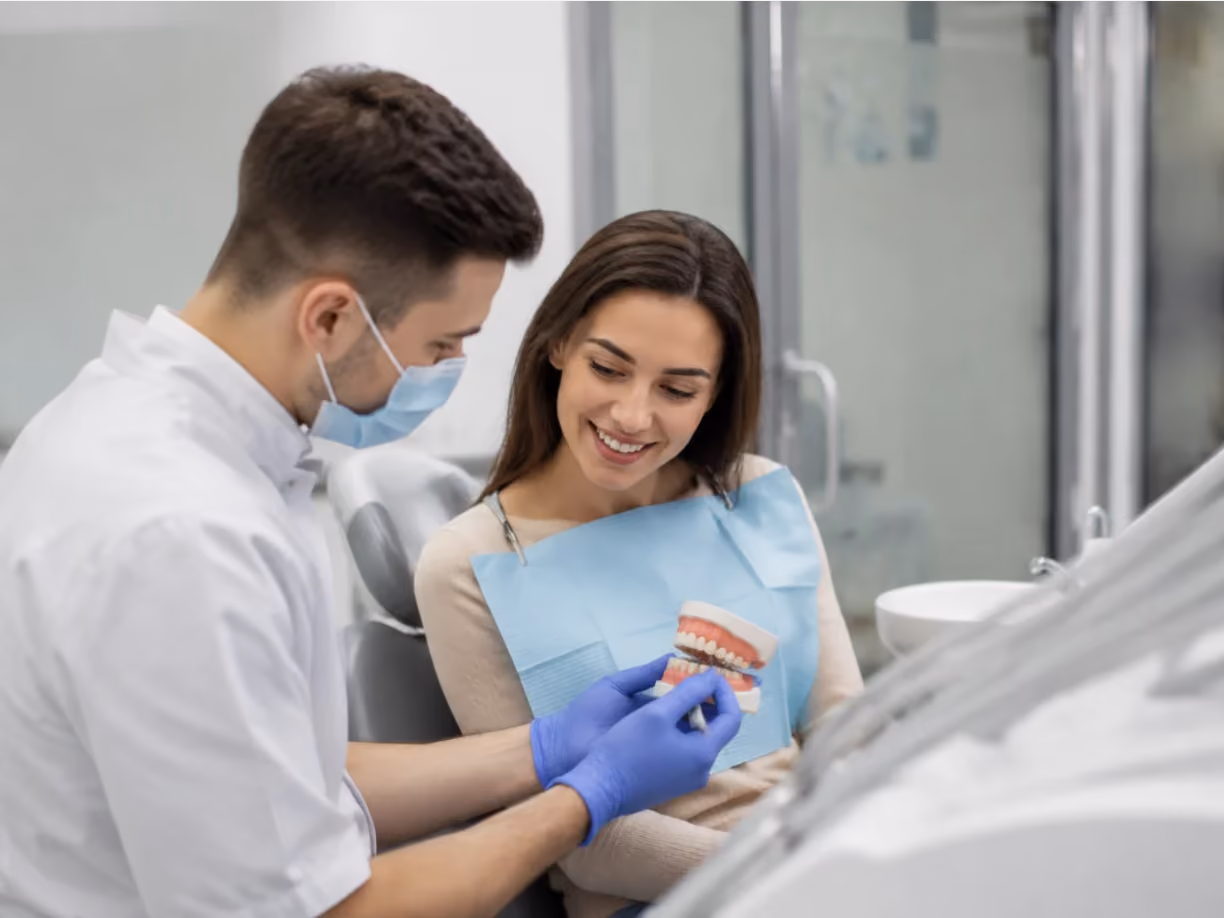 Dentist wearing mask and gloves showing a dental model to a smiling female patient in the clinic.