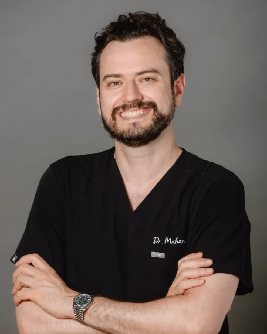 Smiling man with beard wearing black medical scrubs and watch, arms crossed against gray background.