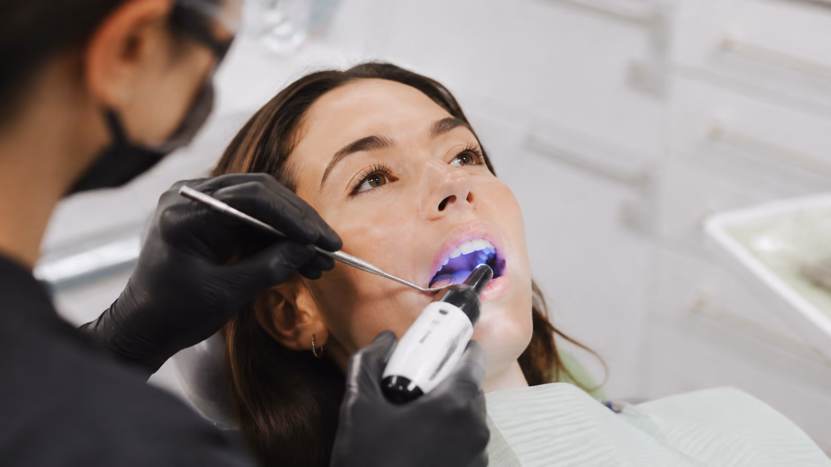 Dentist wearing black gloves using a curing light inside a woman's open mouth during treatment.