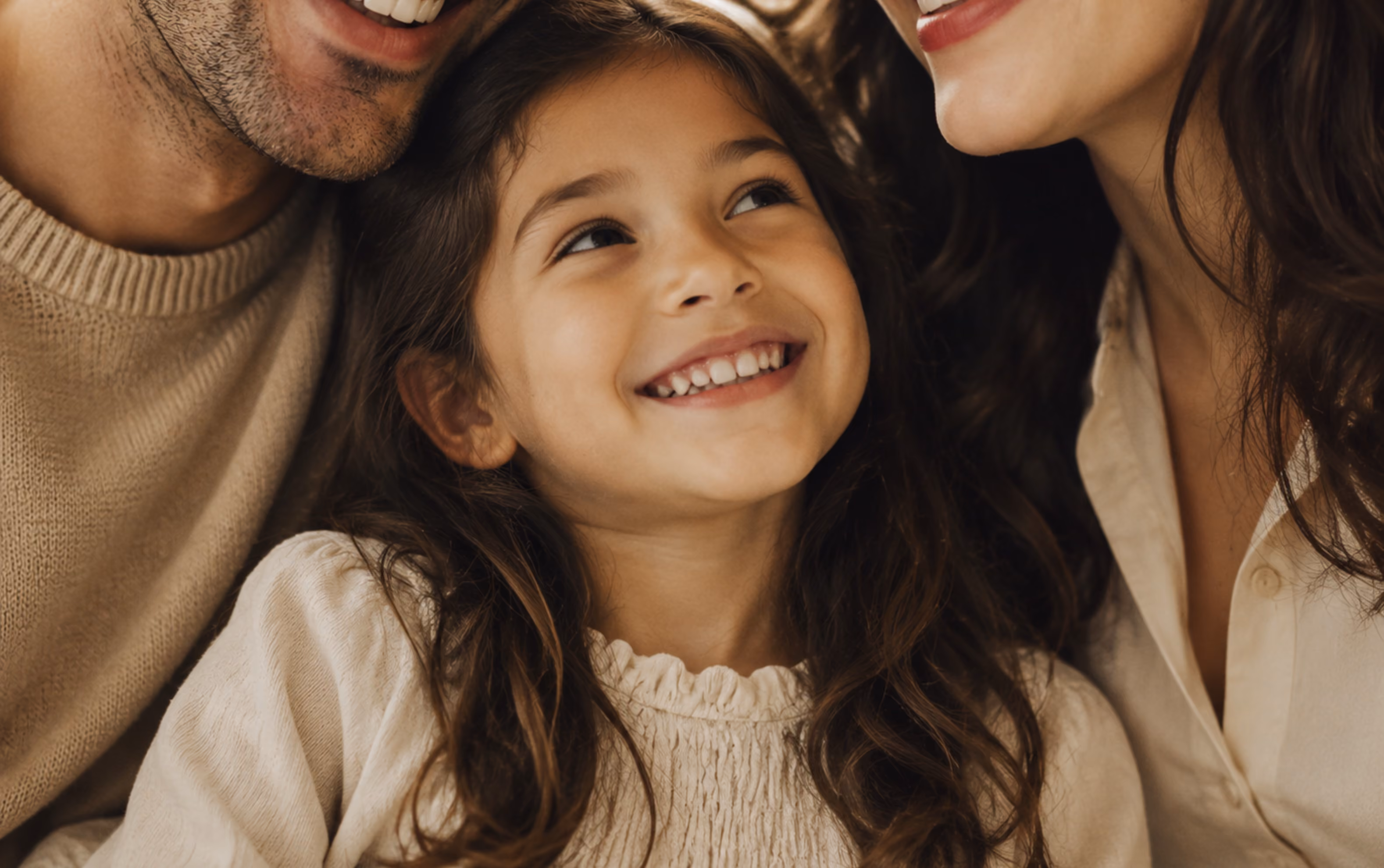 Smiling young girl with wavy hair, looking up between two adults also smiling.