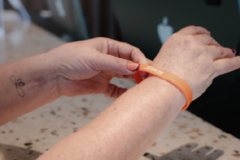 Una persona coloca una pulsera naranja de SFORZA en la muñeca de otra, transmitiendo cercanía, bienestar y sentido de comunidad.