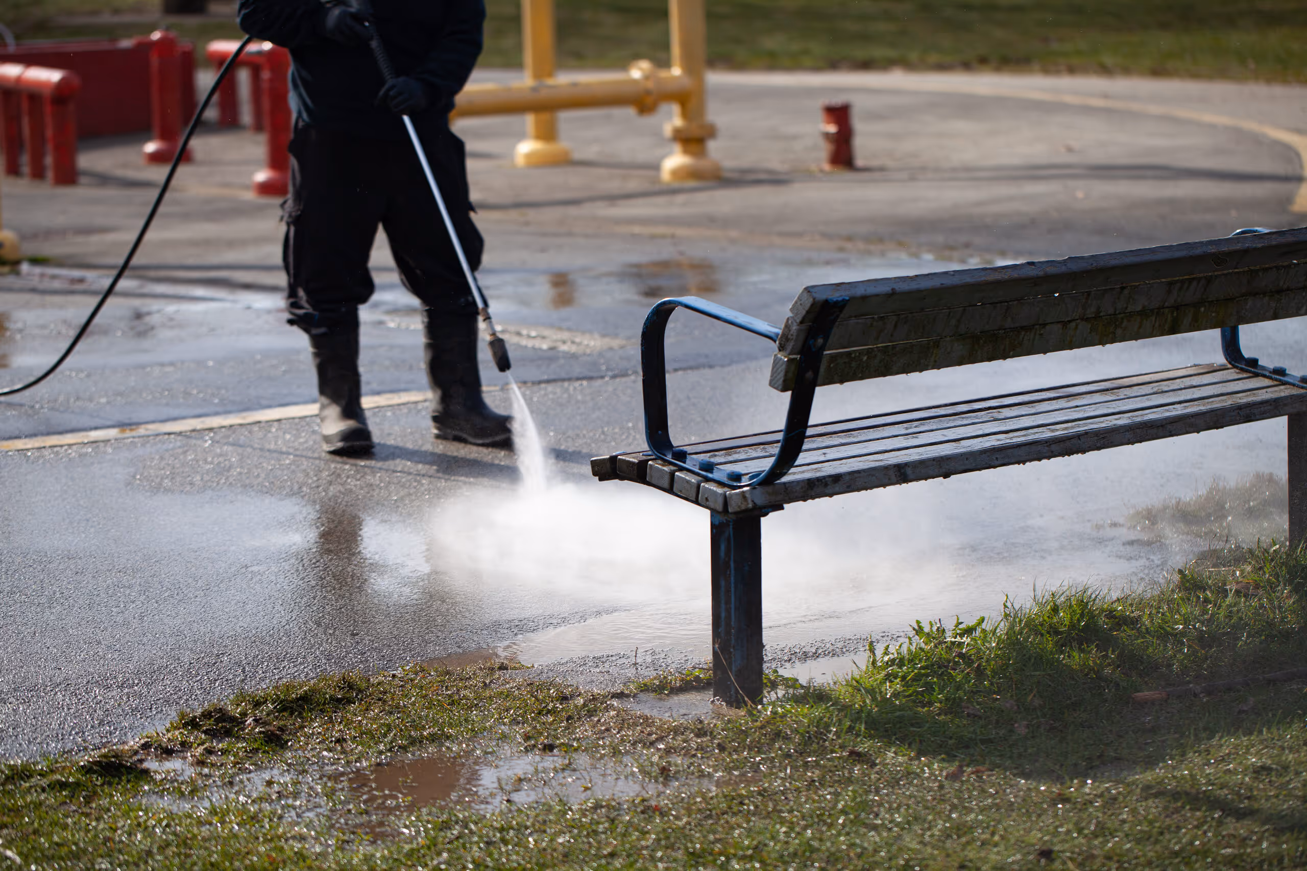 Worker pressure washing a wet playground path near a wooden bench with metal armrests.