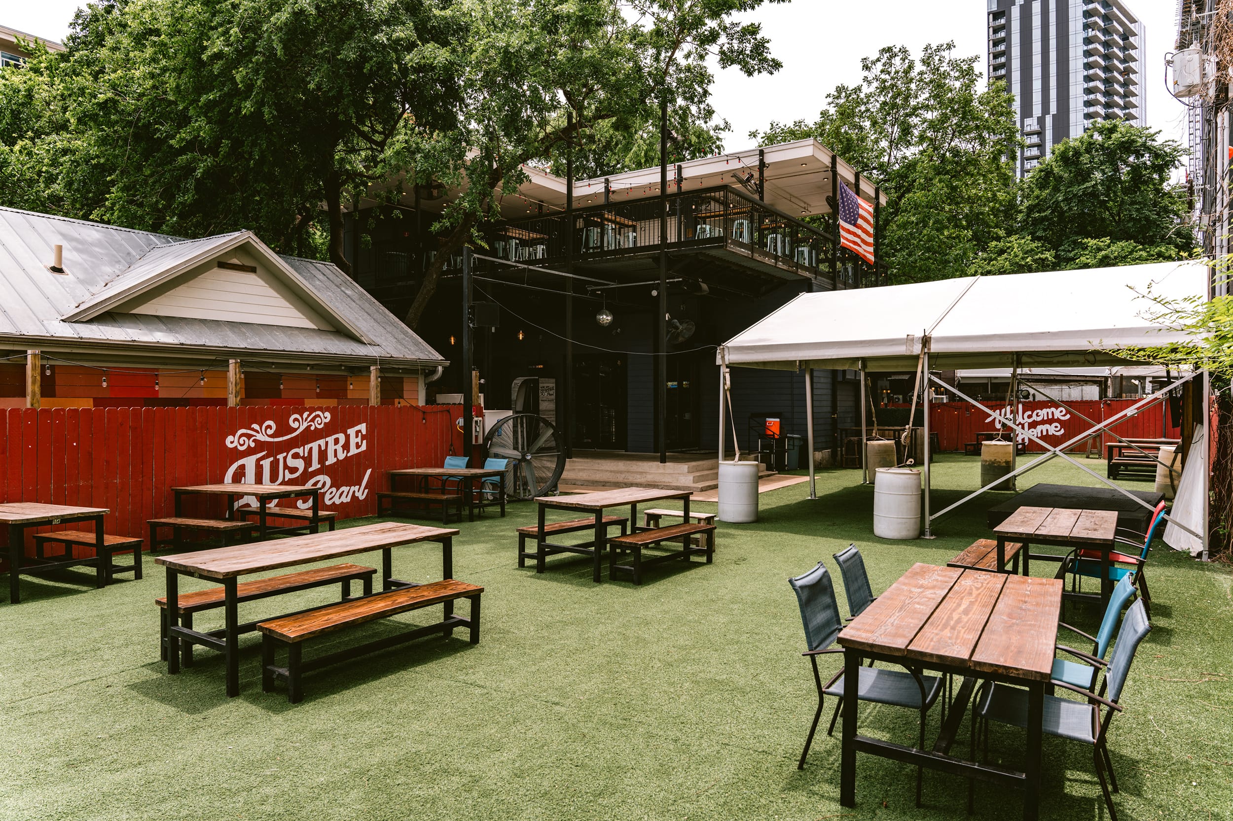 Outdoor patio with wooden picnic tables and benches on artificial grass, a large tree, a red fence with 'Lustre Pearl' painted, a white tent, and a two-story building with an American flag.