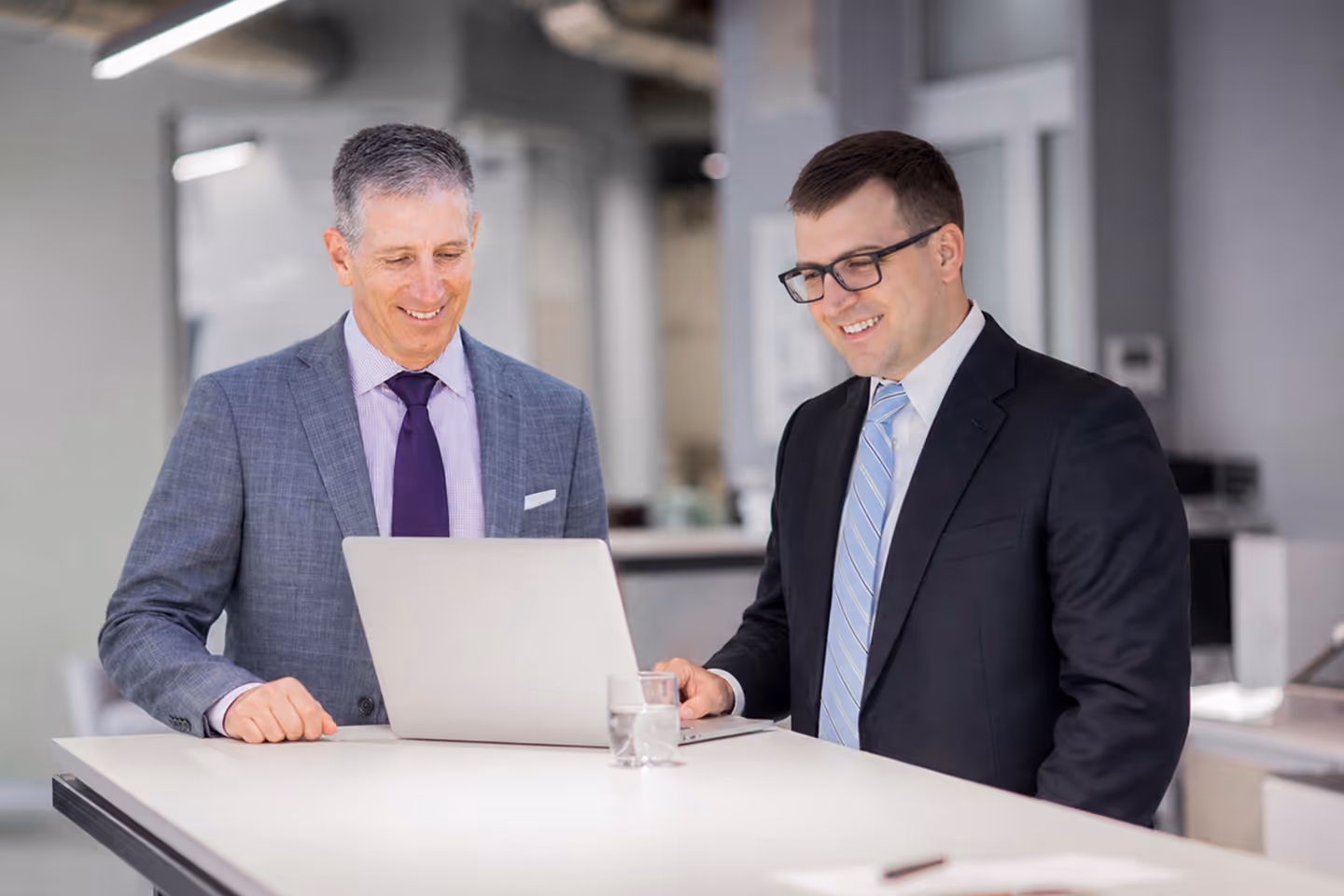 Two businessmen in suits smiling while looking at a laptop on a white office table with a glass of water.