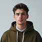 Young man with short brown hair wearing an olive green hoodie against a plain light background.