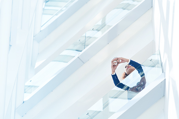 Woman with curly hair wearing business attire, leaning on a glass balcony and looking up while holding a smartphone with both hands in a modern building with white structural beams.