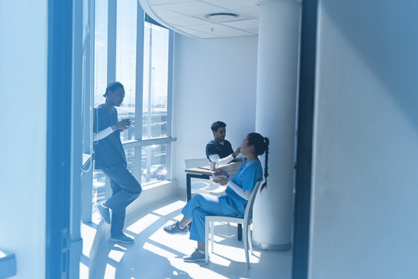 Three healthcare workers in scrubs taking a break and talking in a sunlit hospital hallway with large windows.