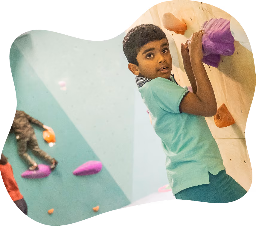 Young boy in a light green shirt climbing an indoor rock wall, holding purple and orange holds, looking at the camera.