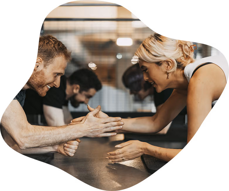 A man and a woman smiling and arm wrestling across a table in a gym setting, with two other people blurred in the background also engaged in arm wrestling.
