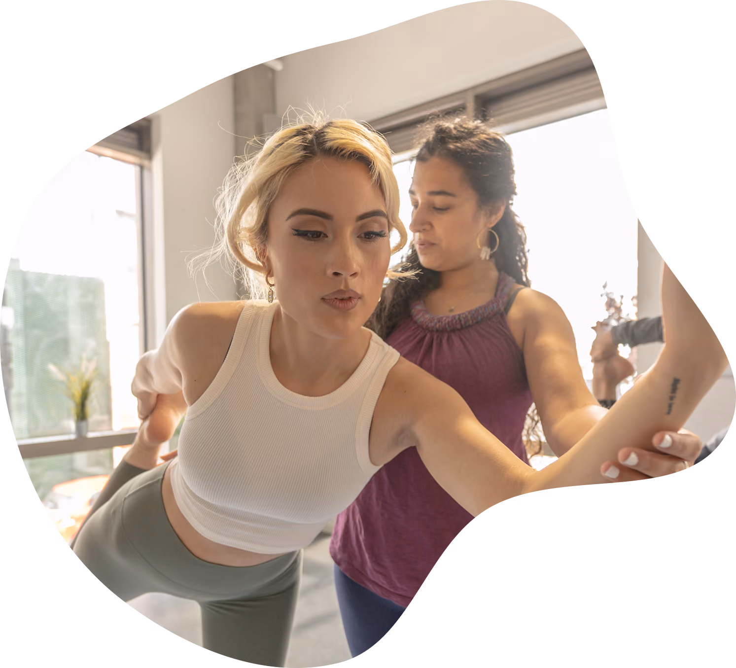 A fitness trainer assists a woman in a white sleeveless top and green leggings with a balance pose indoors near a window.