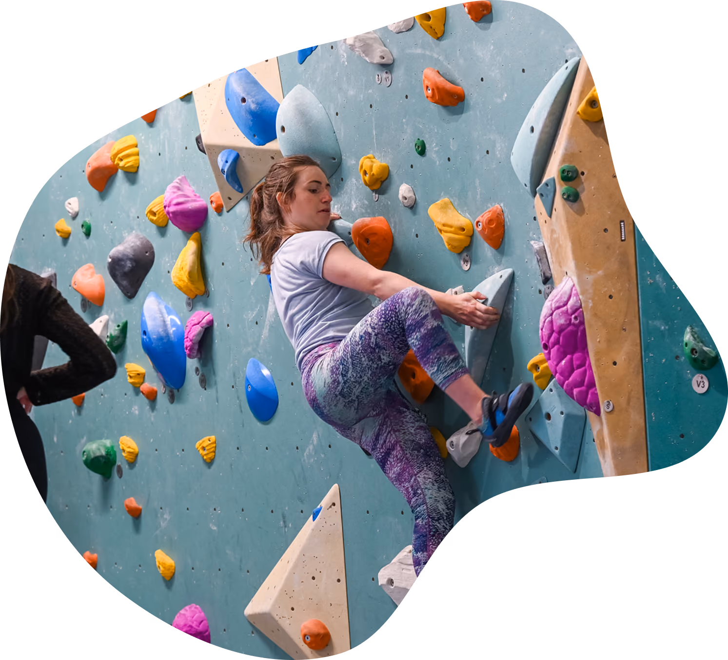 Woman wearing purple patterned leggings and a gray t-shirt climbing an indoor rock climbing wall with colorful holds in various shapes and sizes.