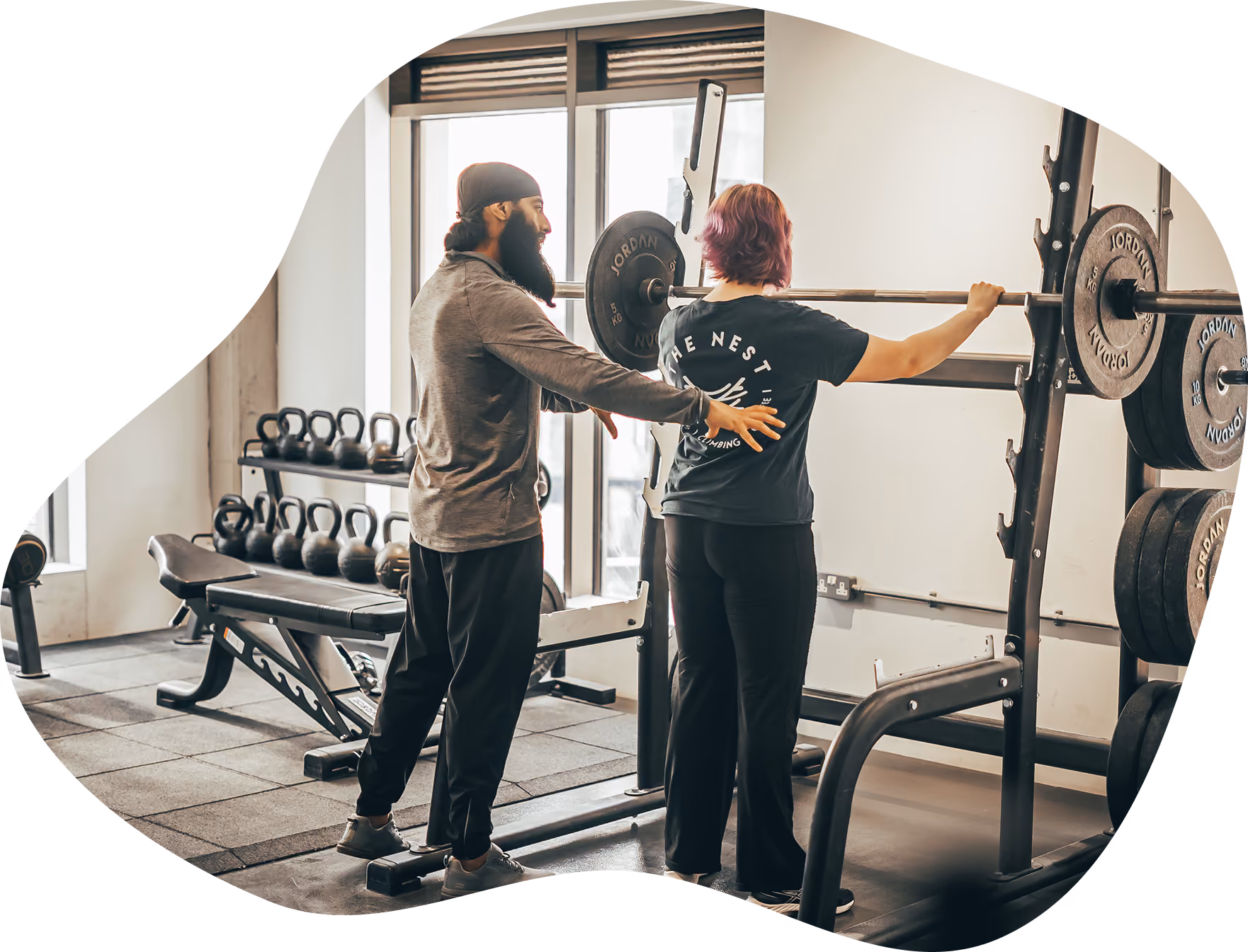 A fitness trainer assists a person lifting a barbell on a squat rack inside a gym with kettlebells and weight plates in the background.