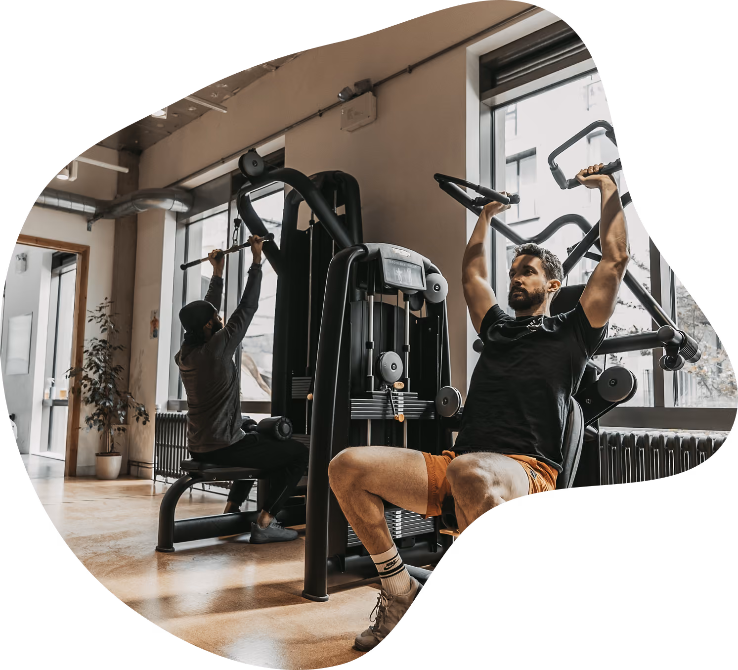 Two men working out in a gym using weight machines near large windows; one man is seated in front focusing on an overhead press exercise, the other is seated behind doing a lat pulldown.