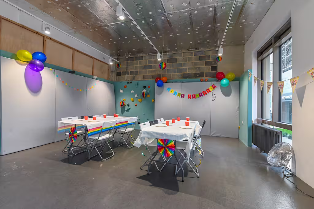 Room decorated for a birthday party with two tables covered in white cloths and decorated with rainbow patterns, colorful balloons on walls and ceiling, orange cups on tables, and a Happy Birthday banner on the wall.