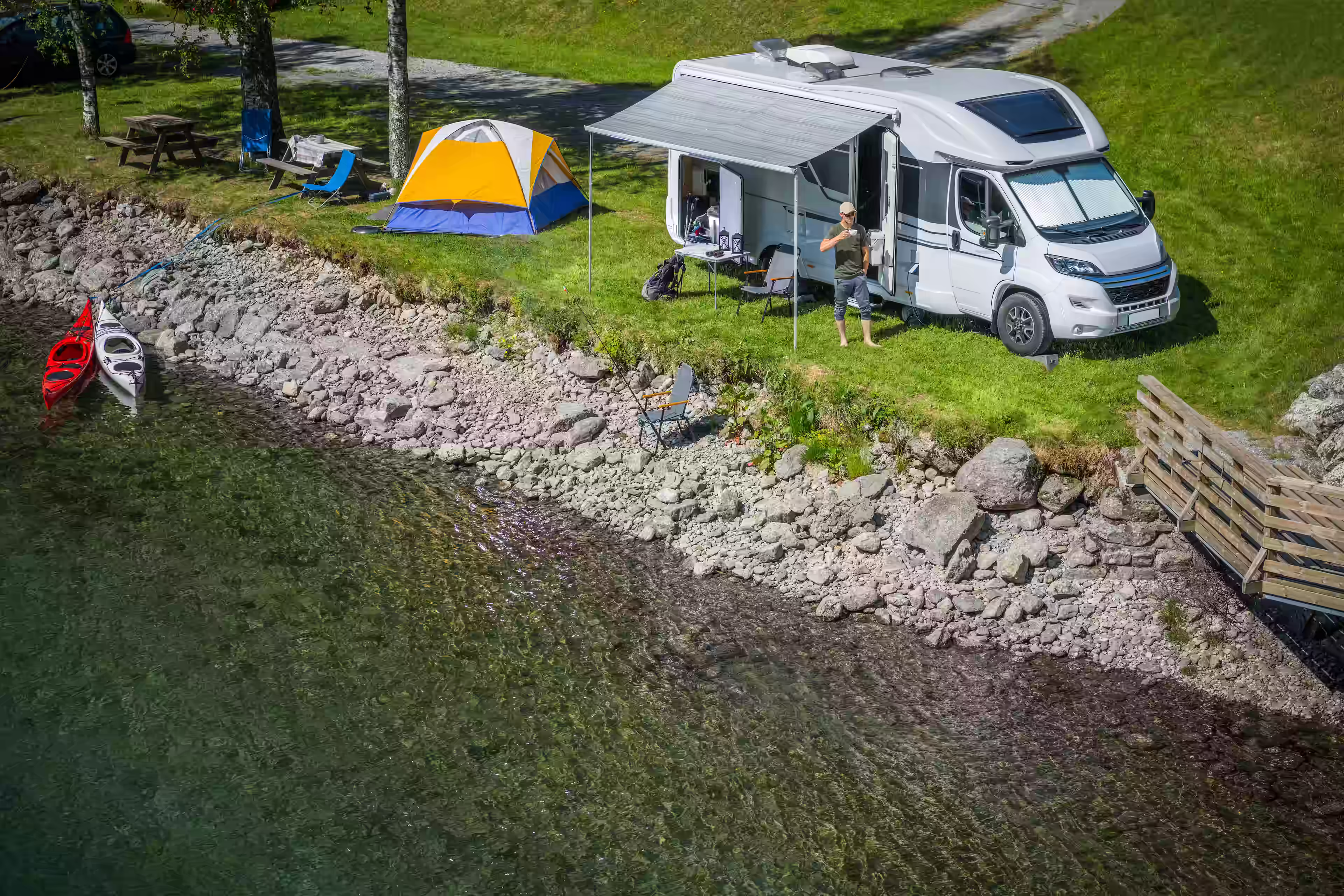 Man camping lakeside with camper van, kayaks, tent, and fishing gear sipping coffee in the morning sun