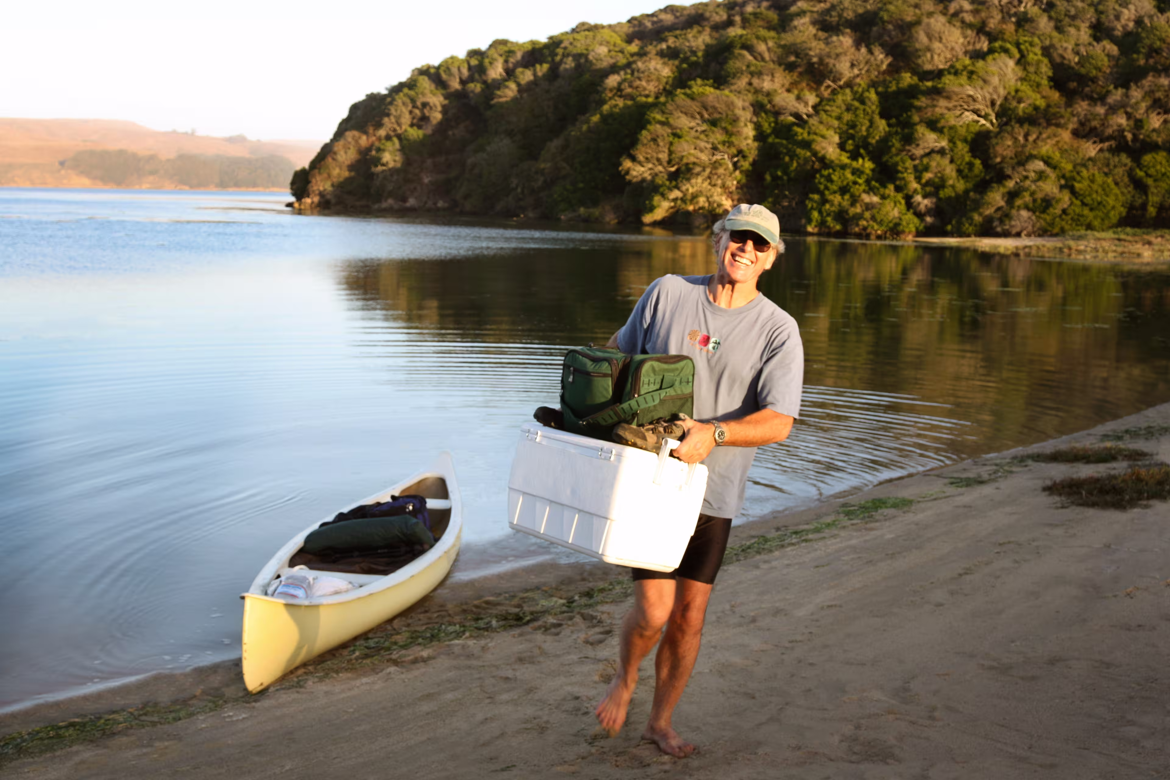 Retiree man carrying a large cooler from his kayak onto a lake front beach