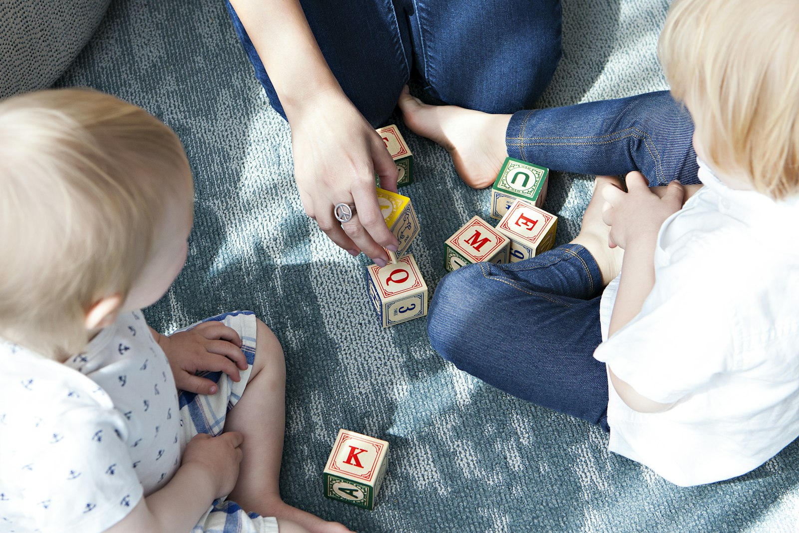 An adult and two toddlers playing with wooden alphabet blocks on a soft rug at home
