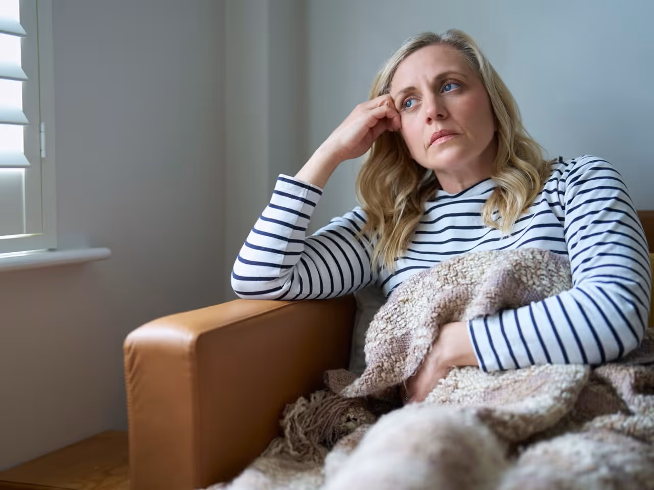 A woman sitting indoors wrapped in a blanket, looking thoughtfully out a window, representing aging, wellness, or emotional health