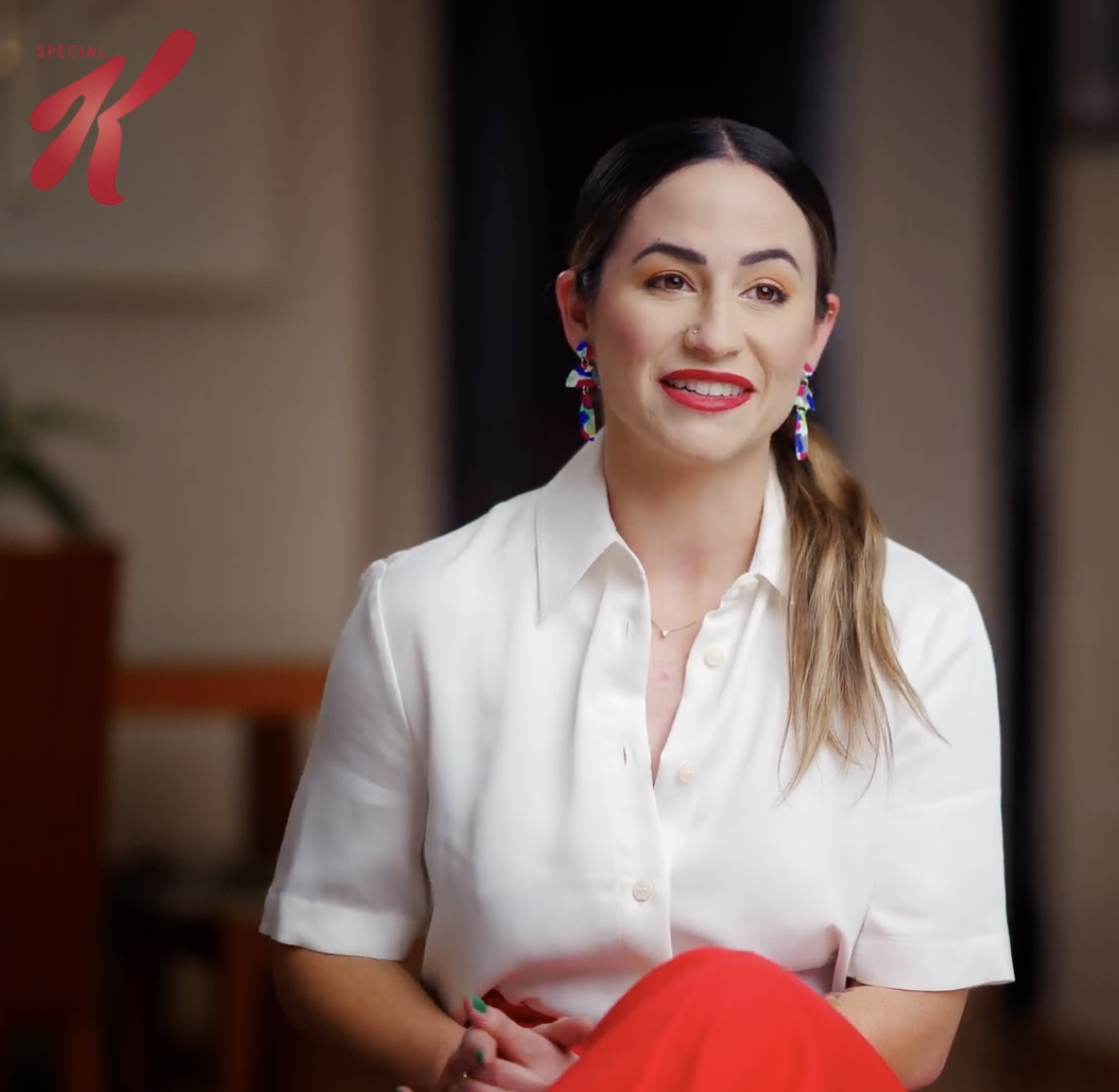 A woman speaking during an interview while seated indoors, wearing a white blouse, colorful earrings, and red pants.