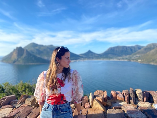 A woman standing at a scenic coastal overlook with mountains and the ocean in the background