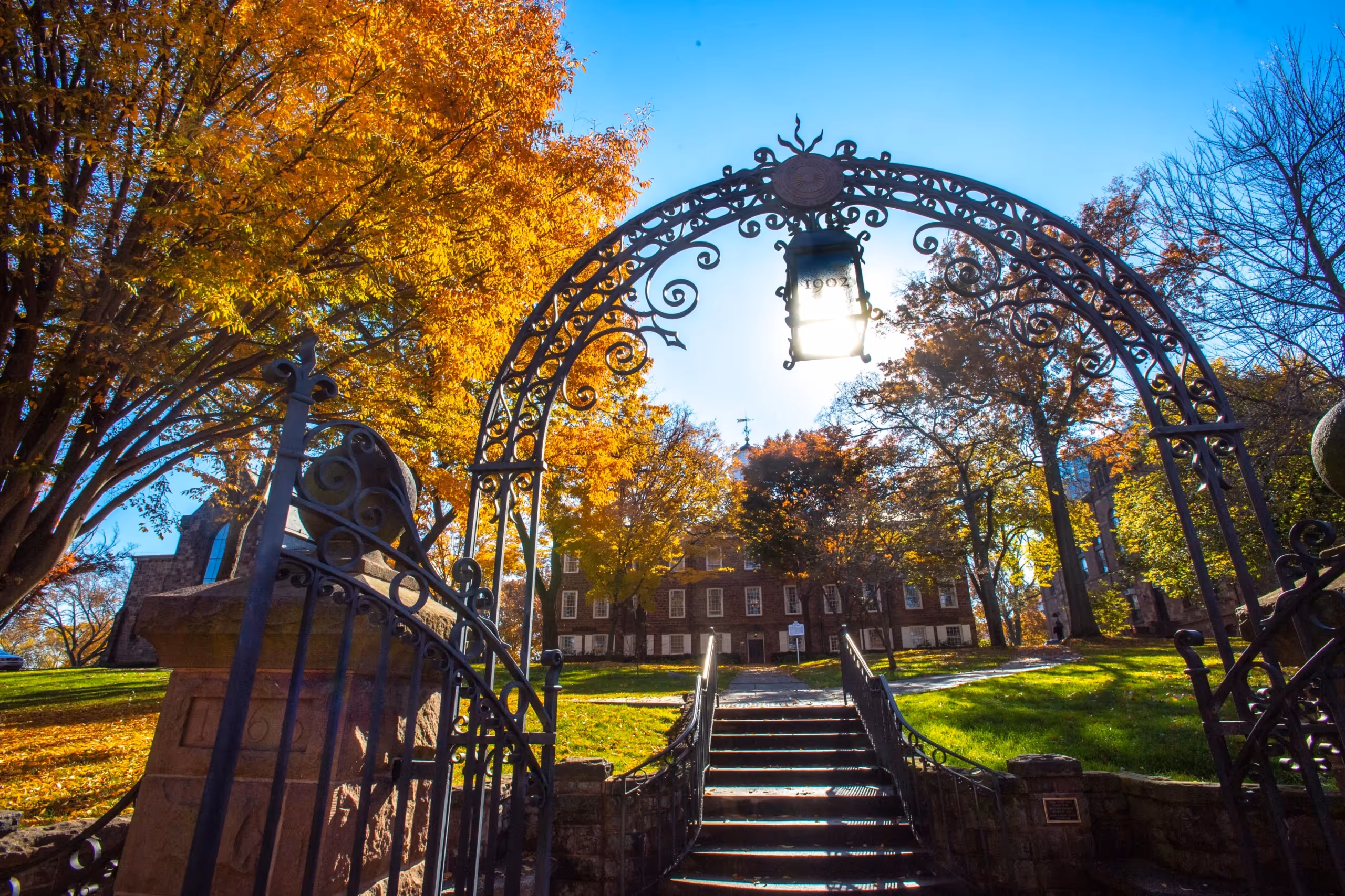 Ornate iron gate with lantern above stairs framed by autumn trees and a historic brick building behind.
