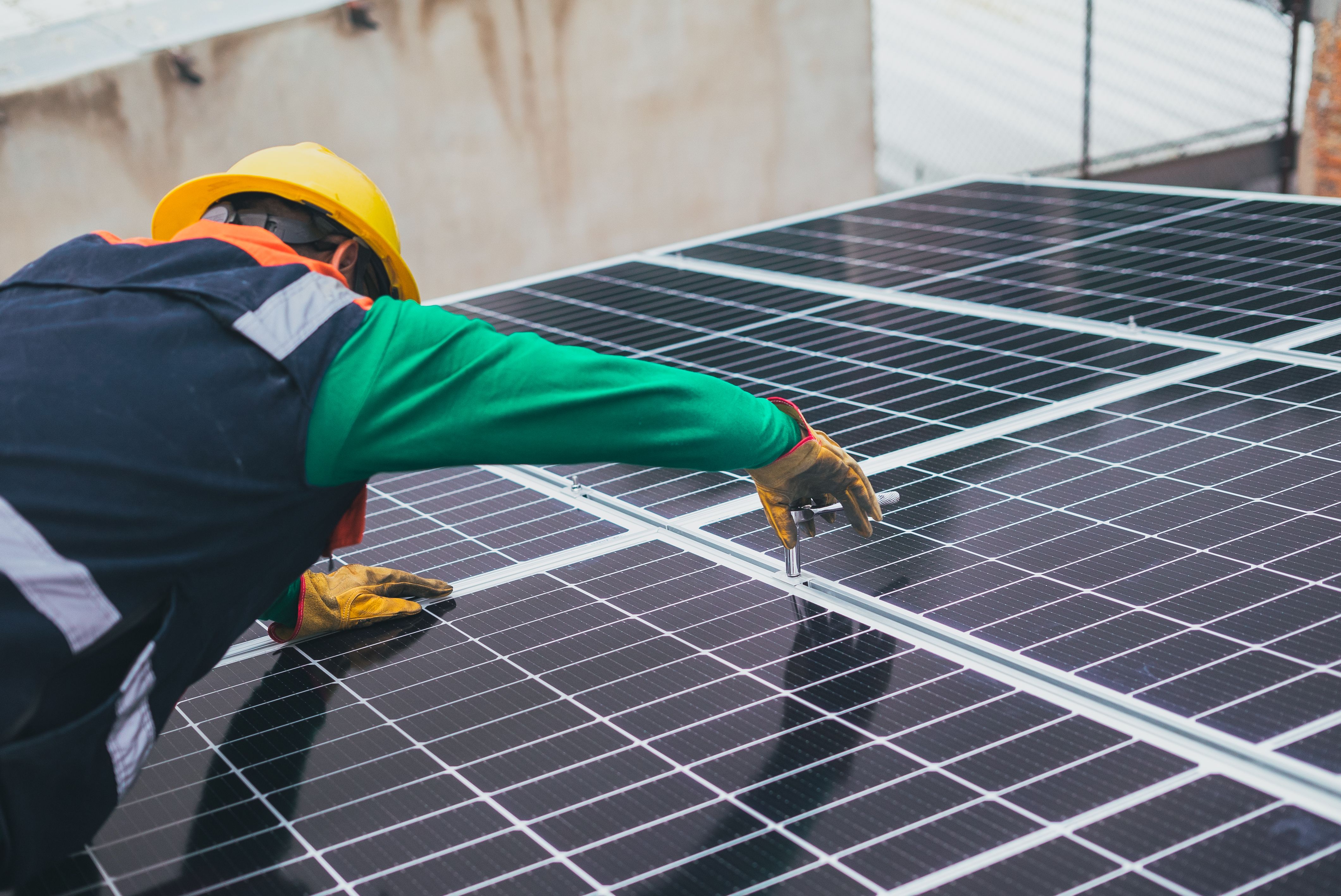 Worker wearing a yellow helmet and gloves tightening bolts on solar panels outdoors.