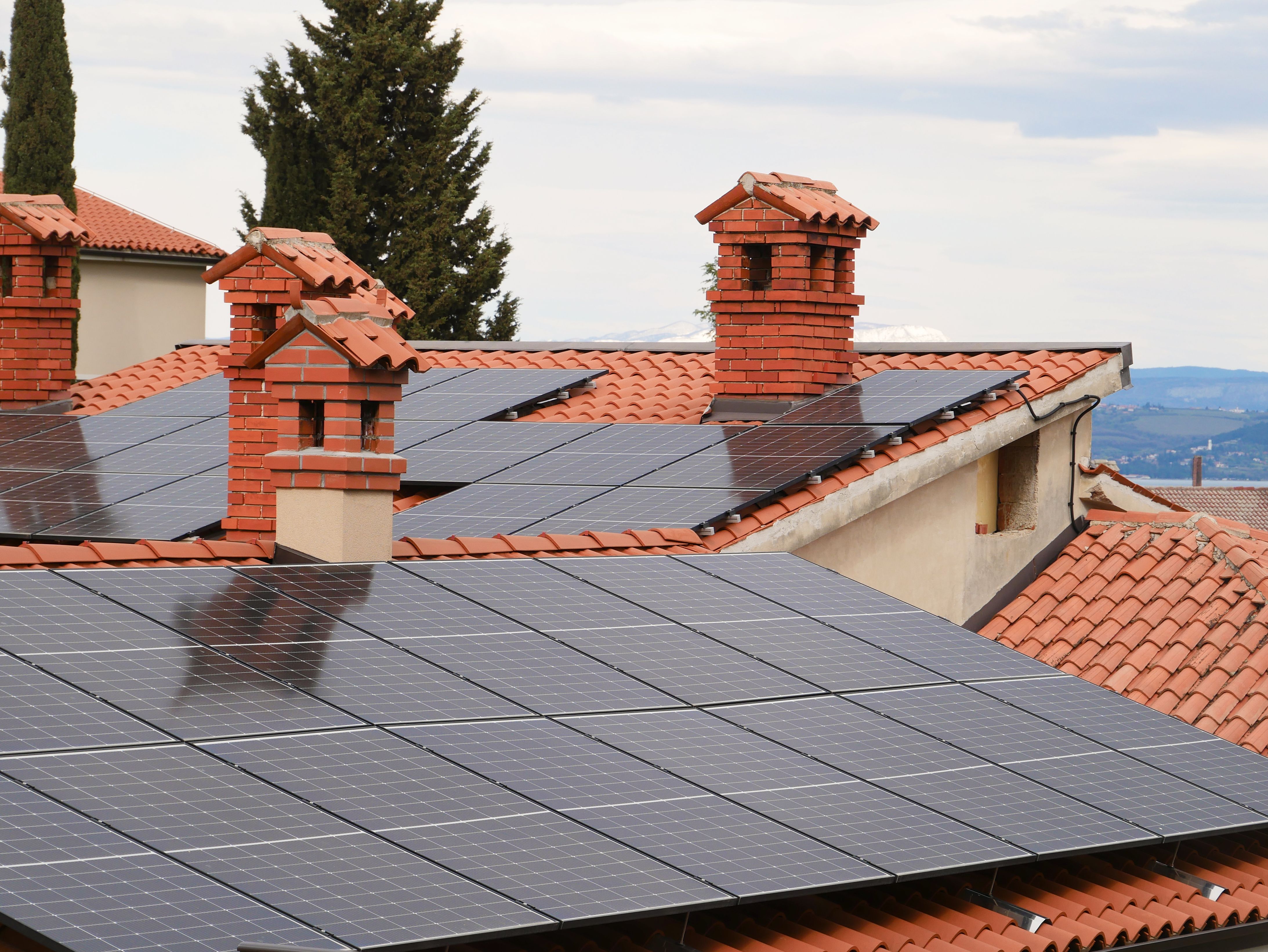 Solar panels installed on red tile roofs with brick chimneys and trees in the background.