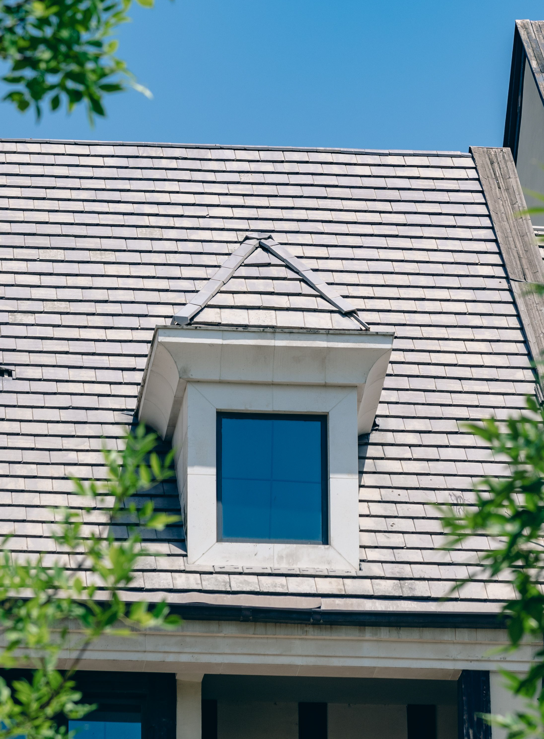 Dormer window with a triangular roof on a gray shingled house roof under clear blue sky.