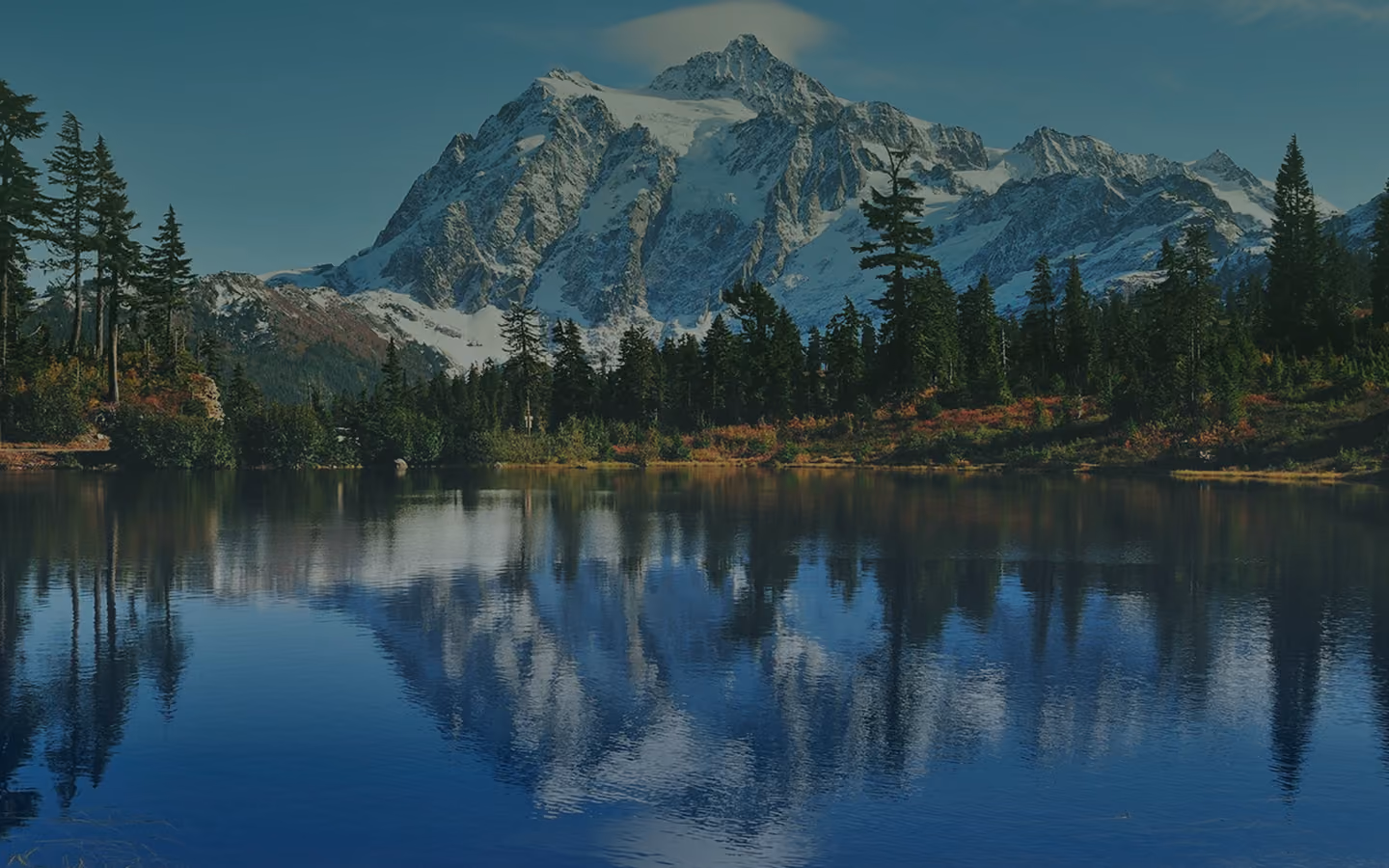 Snow-covered mountain reflected in a calm lake surrounded by evergreen trees under a clear sky.