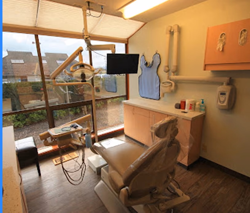 Dental exam room with patient chair, overhead light, monitor, and large windows overlooking houses.