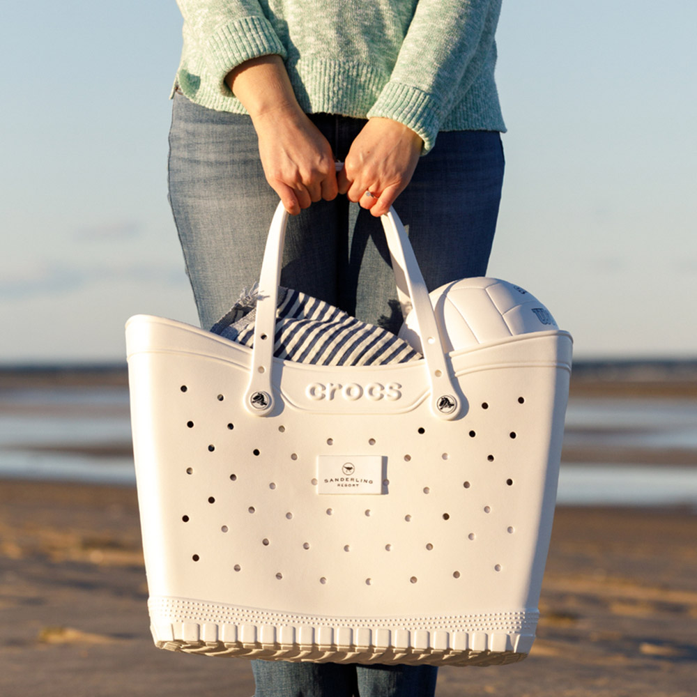 Person carrying a white Crocs tote with a volleyball and striped towel at the beach