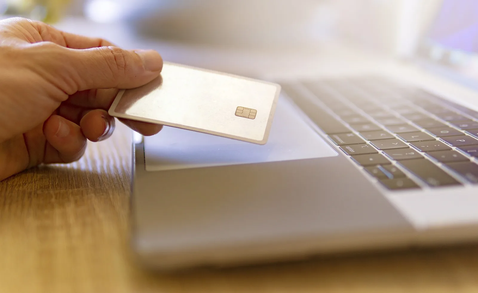 Hand holding a payment card beside an open laptop on a wooden desk