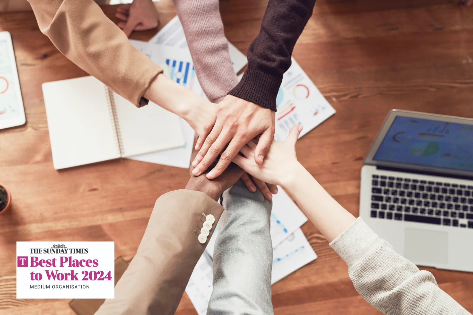 Team members stacking their hands together over a desk, with The Sunday Times Best Places to Work 2024 badge